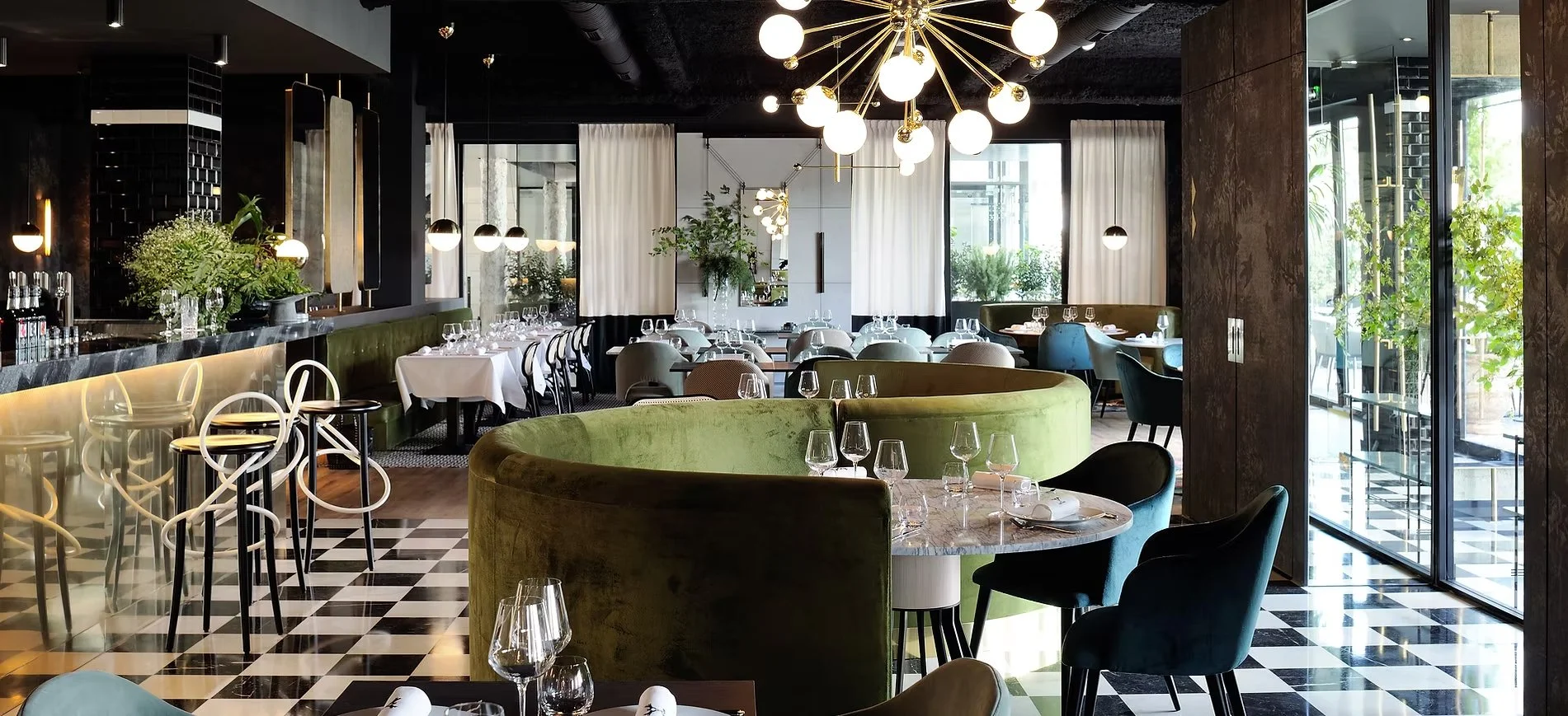 L'intérieur d'un restaurant. Au premier plan un canapé en velour vert, des tables dressées avec des couvert et des verres. Lieu élégant avec une décoration noire, doré, et des matières noble comme le bois et du marbre.