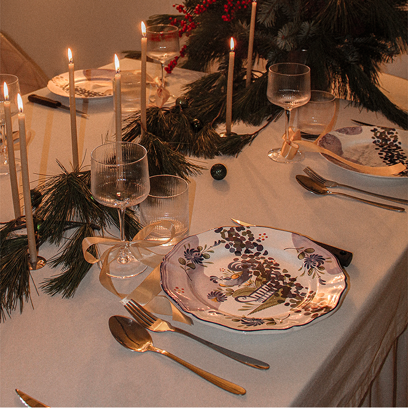Table de fête avec assiettes décoratives, verres, couverts, bougies allumées, et décoration de sapin de Noël en arrière-plan.