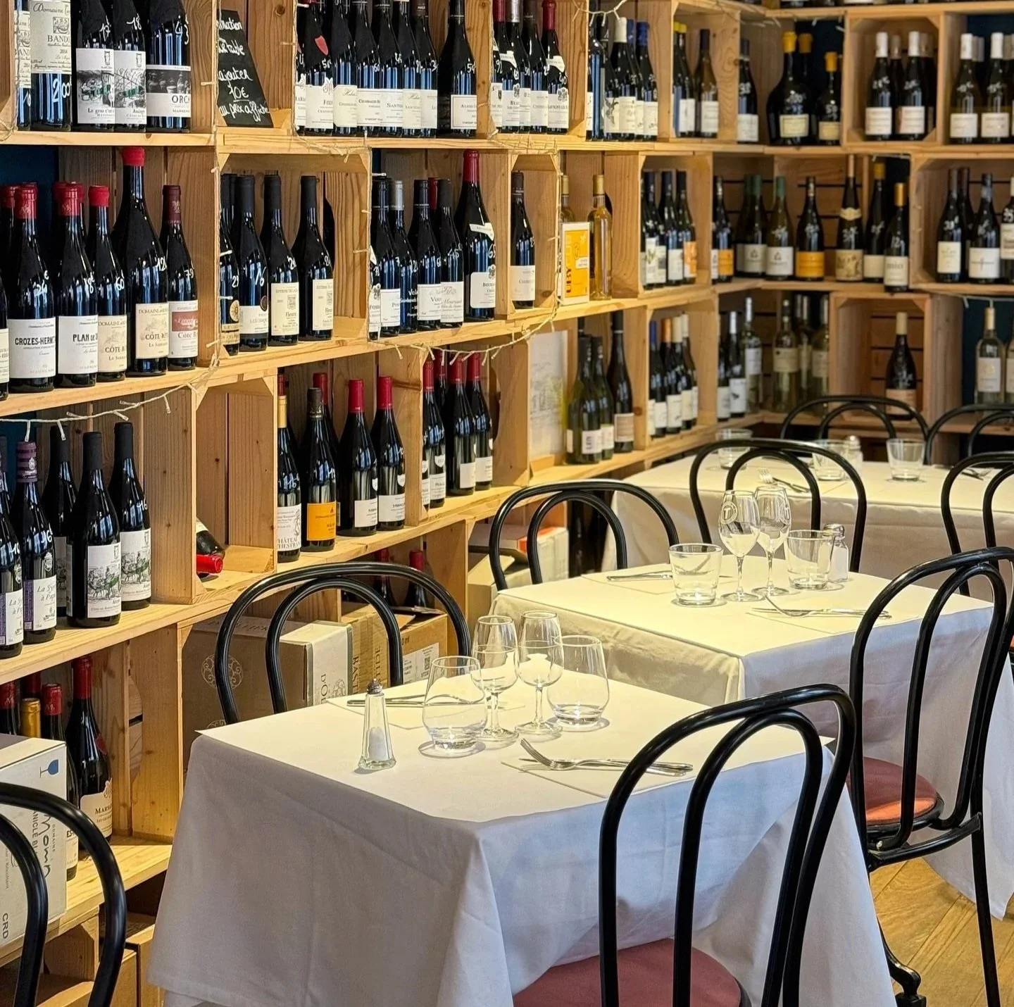 Table dressées avec verres et couverts, chaises en bois. Derrière, un mur avec des étagères remplies de bouteilles de vin.
