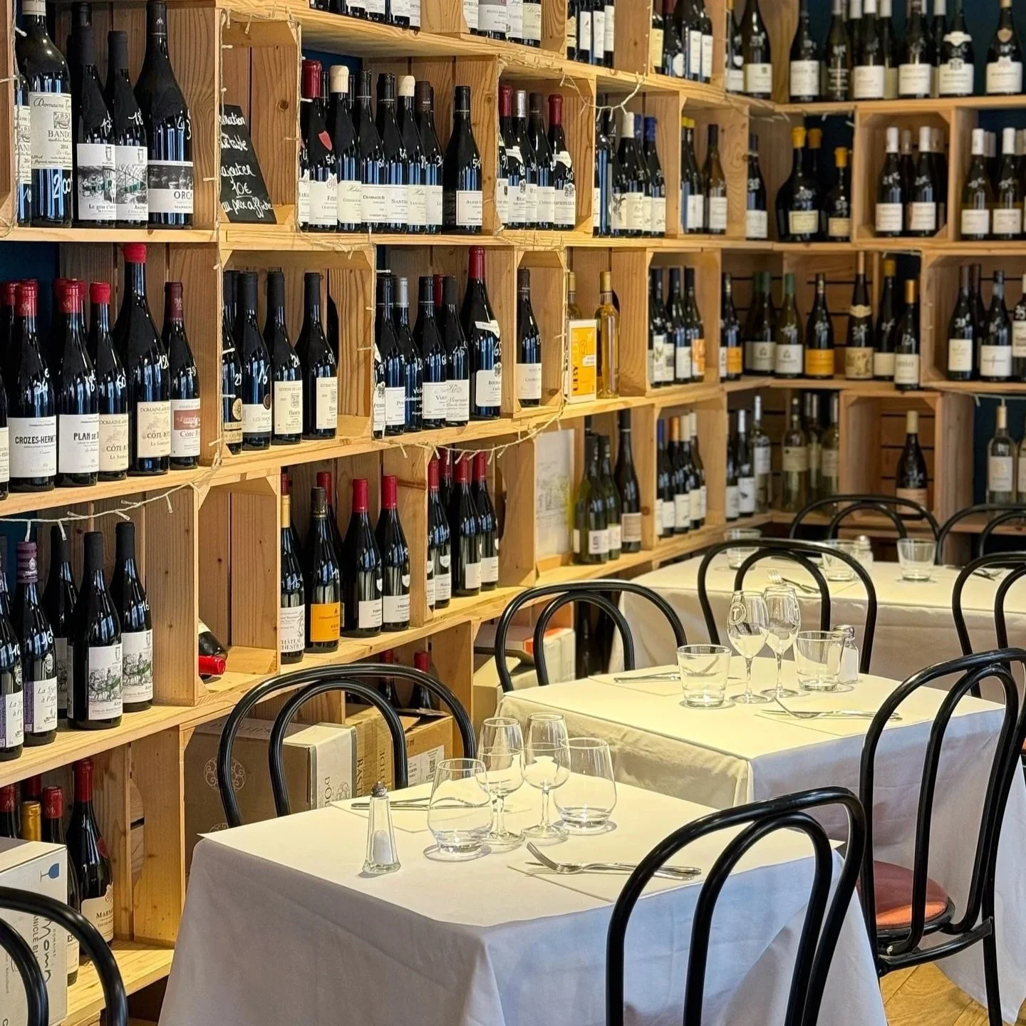 Table dressées avec verres et couverts, chaises en bois. Derrière, un mur avec des étagères remplies de bouteilles de vin.