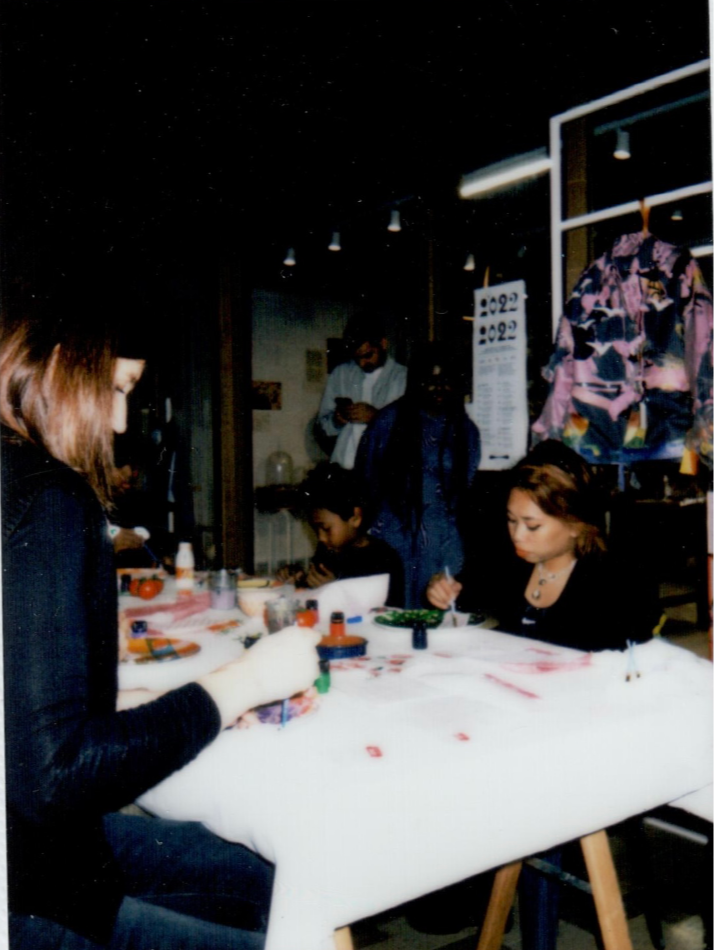 Groupe de personnes, dont des enfants, qui s'engagent dans une activité artistique autour d'une table avec de la peinture. Décor intérieur avec vêtements exposés.