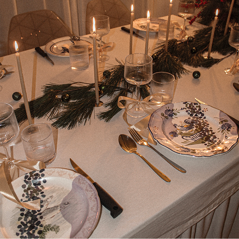 Table de dîner décorée avec des bougies allumées, des branches de pin, des boules de Noël, des assiettes à motifs de saison, des verres à vin, des couteaux et fourchettes en or, et des rubans en satin
