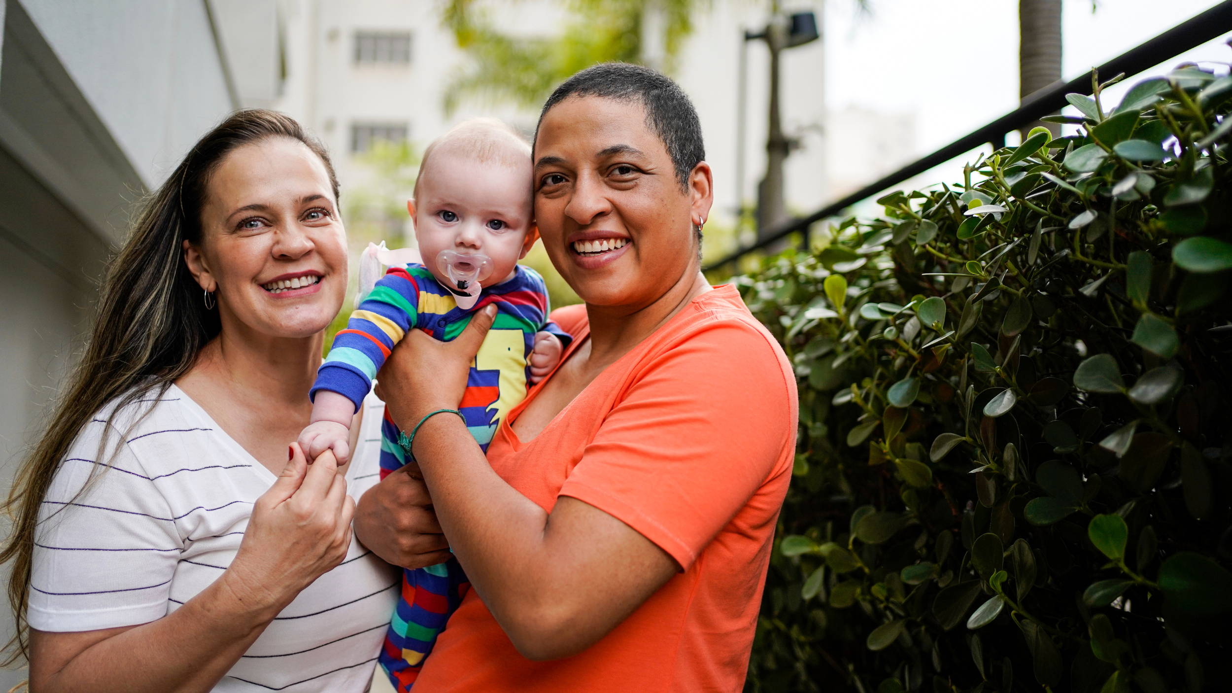 Two mothers smiling while holding their baby outdoors, representing family support, caregiving, and financial planning for young families.