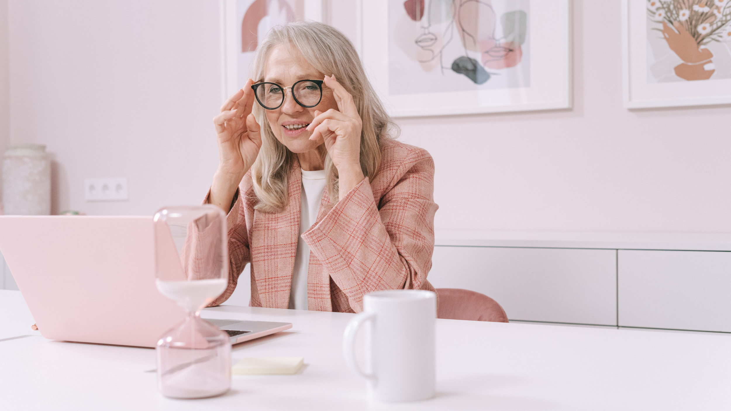 Older woman wearing glasses smiling during a video call on her laptop, seated at a desk with a coffee mug and hourglass in a home office.