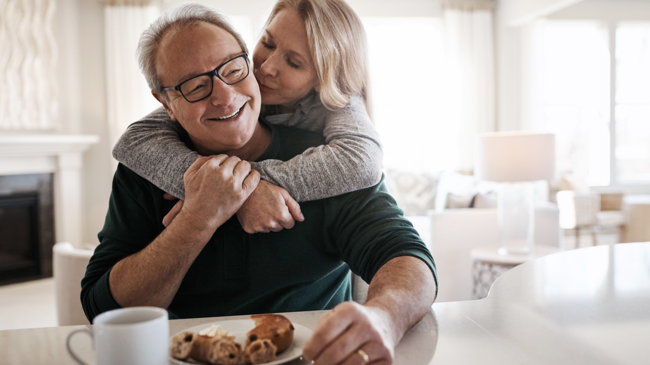 Smiling older couple embracing at kitchen table over coffee and pastries, enjoying peace of mind from retirement planning