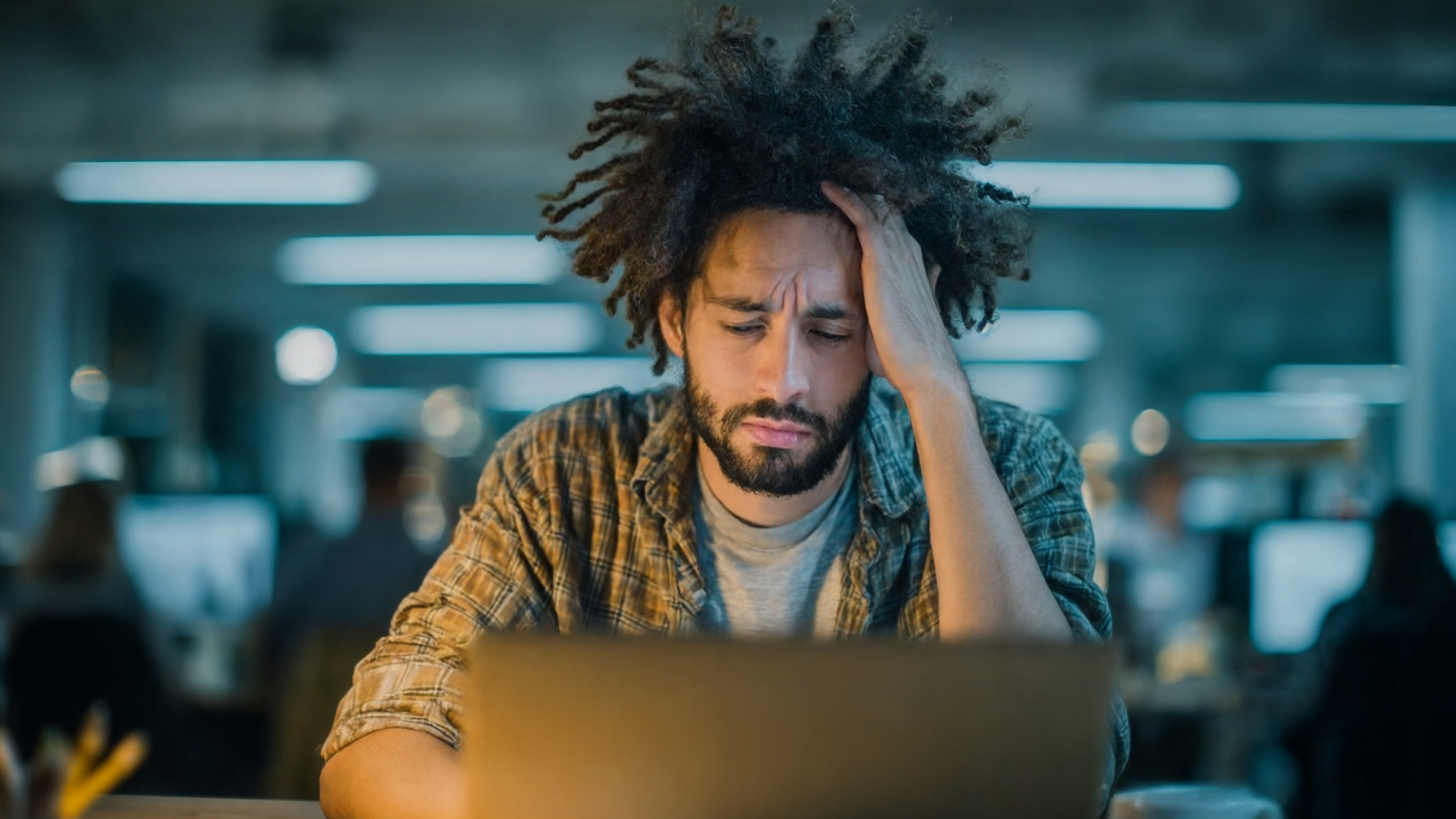 Young man working late on a laptop in an office, holding his head in frustration while dealing with financial stress or work pressure.