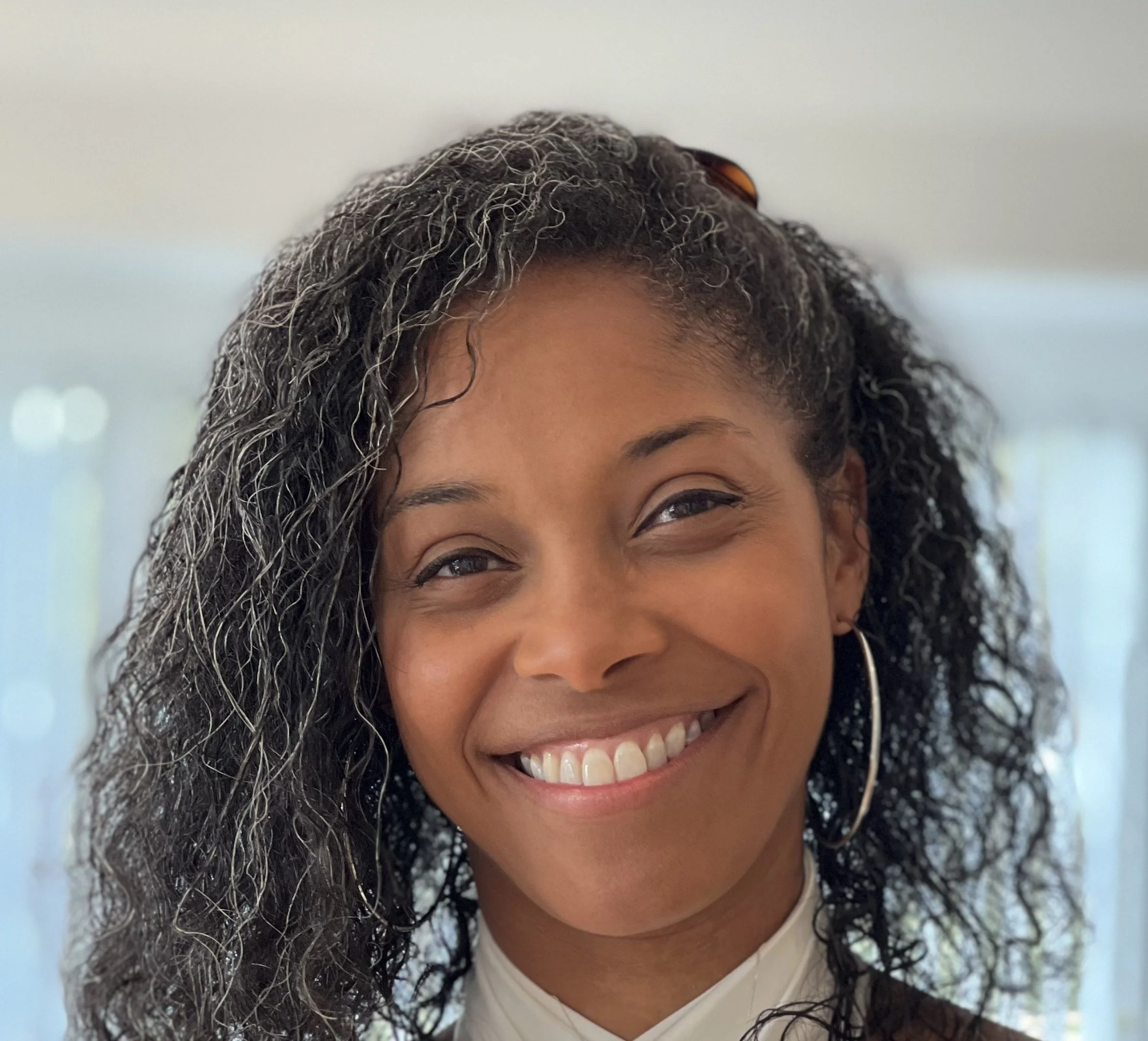 A smiling woman with curly black and gray hair, wearing hoop earrings and a white top.