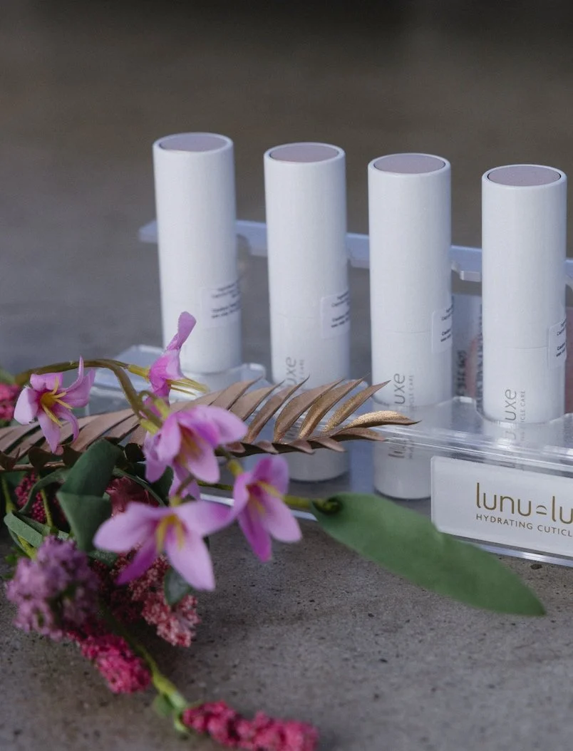 Four white cylindrical containers labeled 'Luxe' in a clear display case with a label reading 'Lunu Luu Hydrating Cuticle' on the front, decorated with pink and purple flowers and gold leaves on a gray surface.