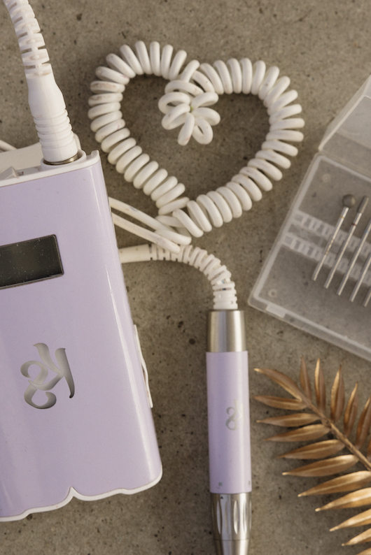 A coiled white curling iron cord forming a heart shape, connected to a purple and silver curling iron, with nail drill bits and golden leaves nearby on a gray surface.