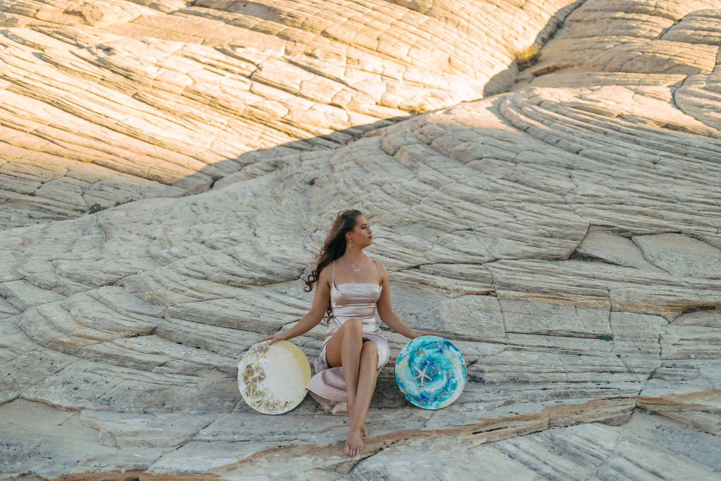 A woman in a beige dress sitting on layered rock formations, in St George, Utah holding two luxury abstract art paintings in blue and gold, with natural elements.