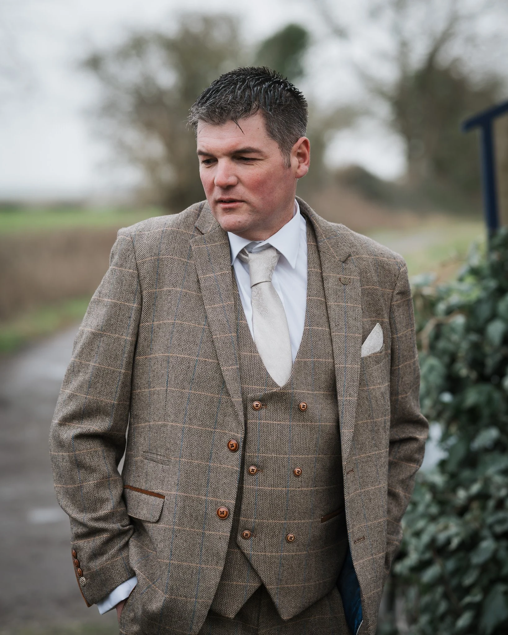 A man in a brown plaid suit with a cream vest and white shirt standing outdoors with blurred trees and greenery in the background.