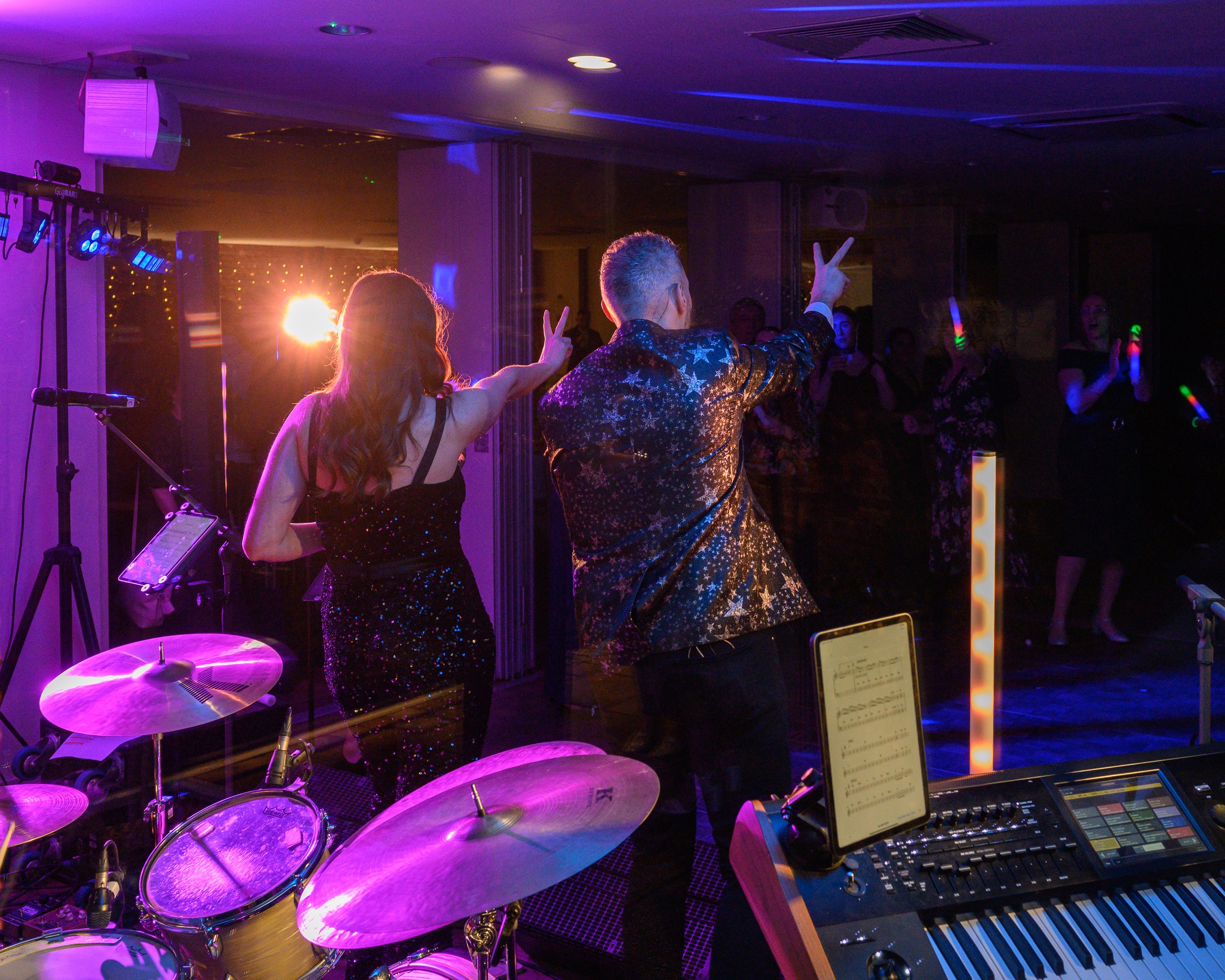 People dancing at a party with colorful lights and a nearby drum set and keyboard.