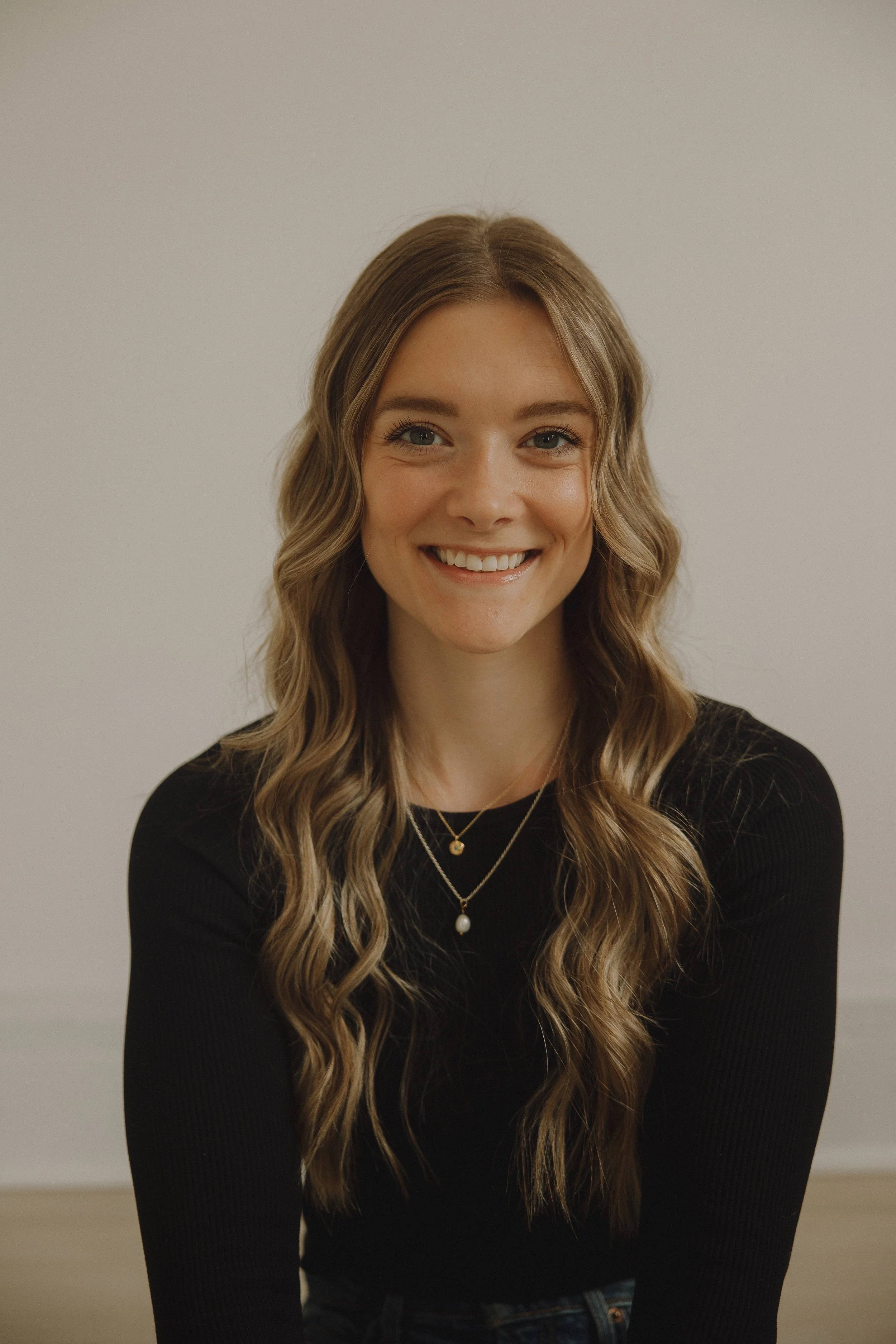 A young woman with long, wavy, light brown hair smiling at the camera. She is wearing a black top and layered necklaces with a pearl pendant. The background is plain and off-white.