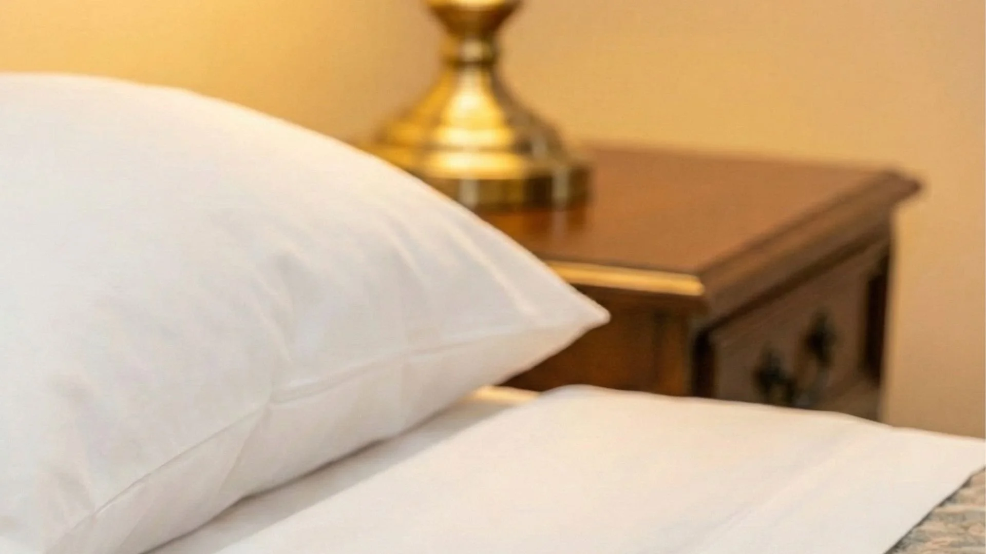 Close-up of a white pillow on a bed with a wooden nightstand and a brass lamp in the background.