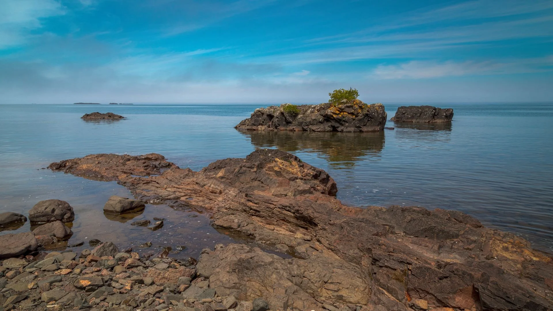 Rocky shoreline with large rocks and calm water, with small islands and a partly cloudy sky in the background.