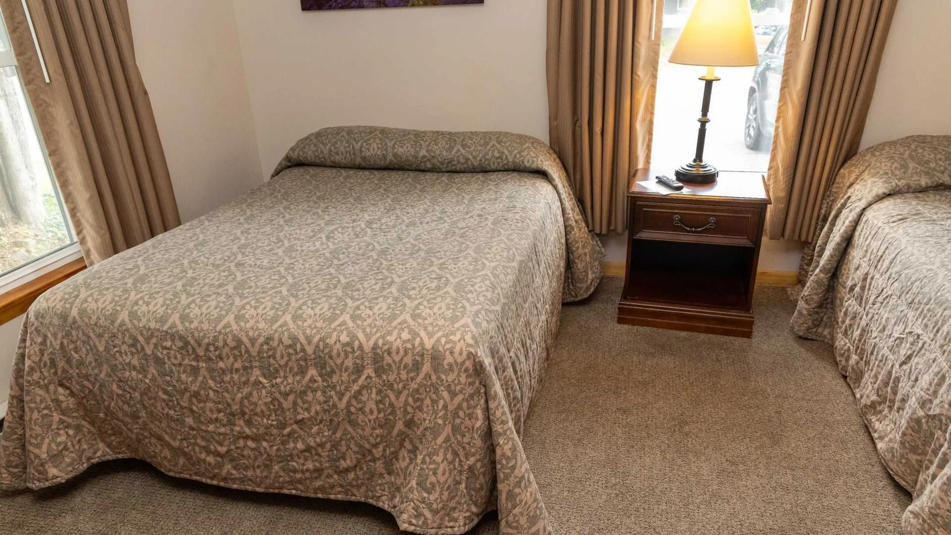 Two single beds with matching bedspreads. A nightstand with a lamp and remote control is between the beds. There are windows with curtains on both sides of the nightstand, and a carpeted floor.