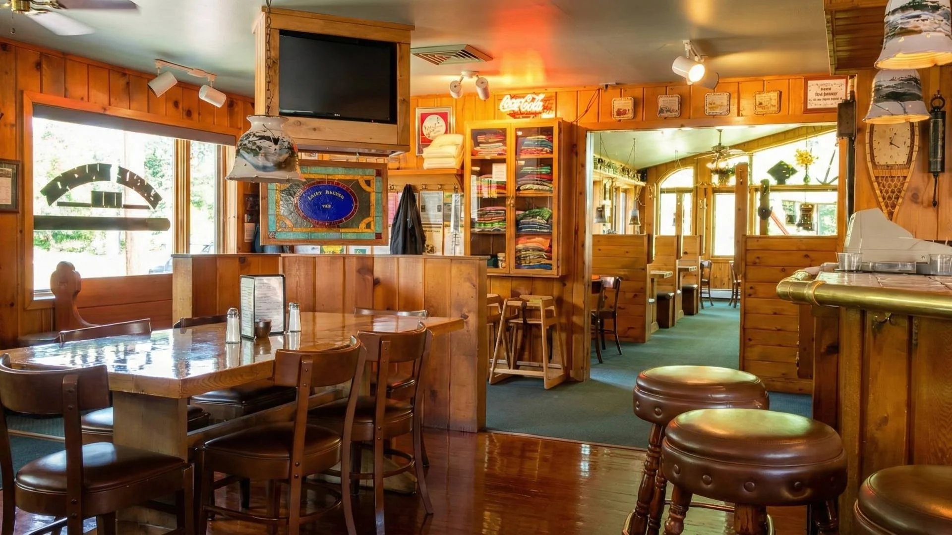 Inside of a cozy wooden restaurant with natural light, featuring a dining table with chairs, a bar counter, and decor such as framed pictures, a neon Coca-Cola sign, and vintage signs on the walls.