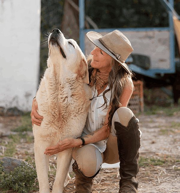 A woman in outdoor clothing and a wide-brimmed hat kneels and hugs a large, fluffy dog that is tilting its head back. The woman is smiling and looking affectionately at the dog.