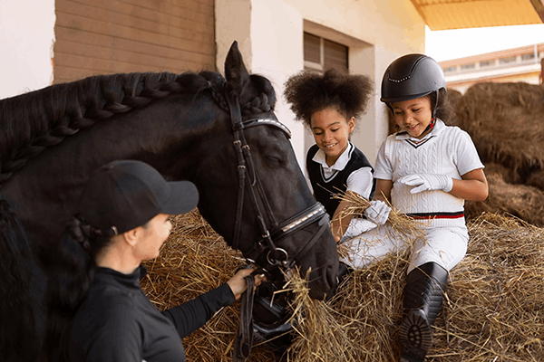 Two young girls riding a horse, one of them wearing a helmet, in a barn with hay, while an adult woman adjusts the horse's bridle.