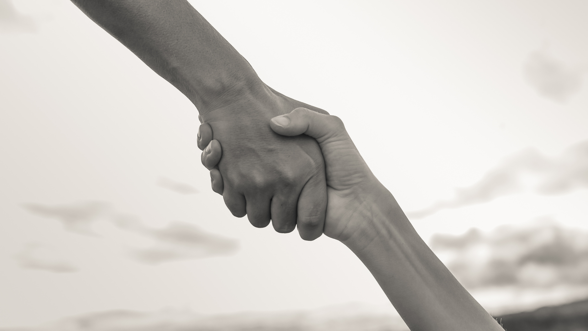 A grayscale photo of two hands shaking hands against a cloudy sky background.