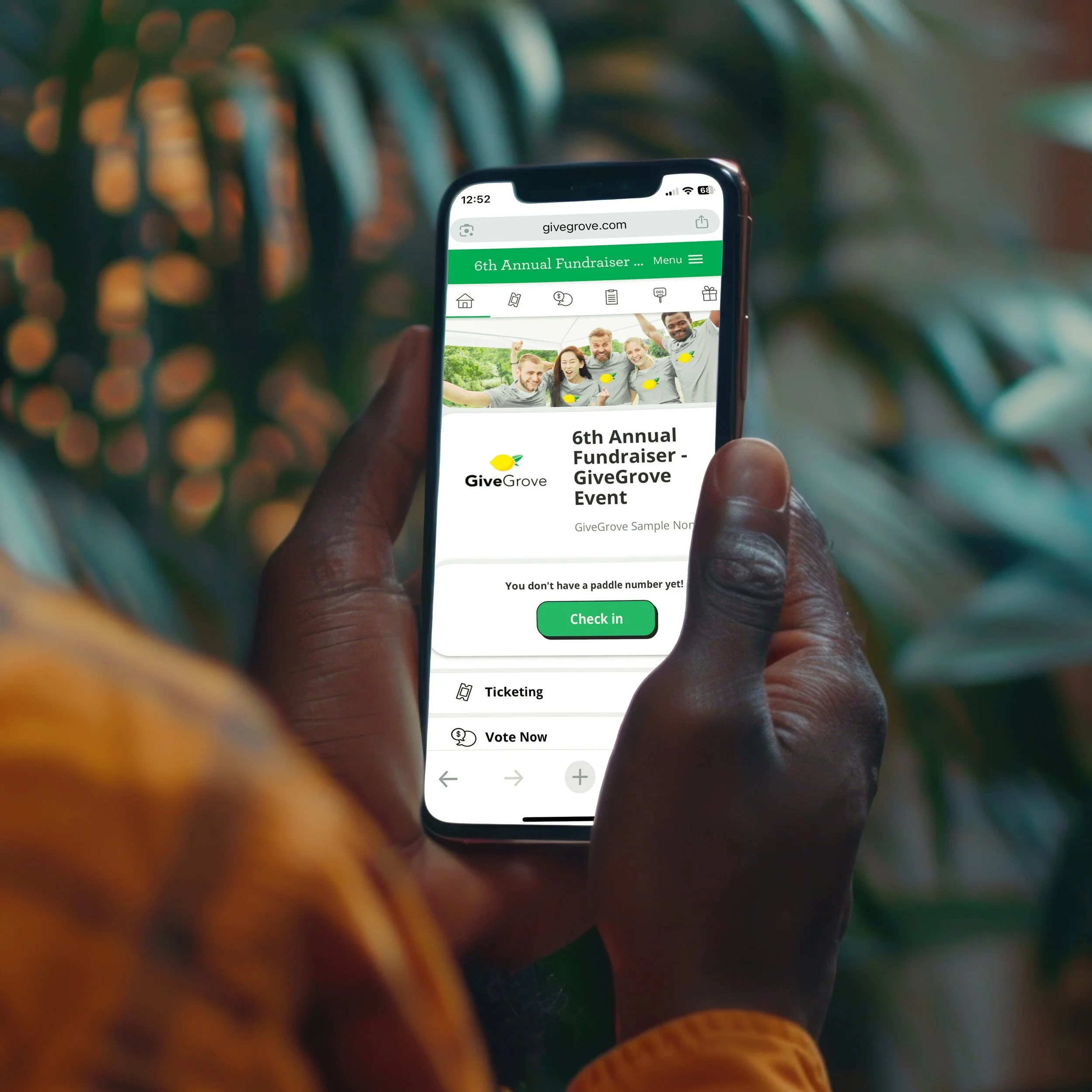 Close-up of a person holding a smartphone displaying a webpage for the 6th Annual Fundraiser Event for GiveGrove, with a green header, a photo of smiling people, and options for ticketing and voting.