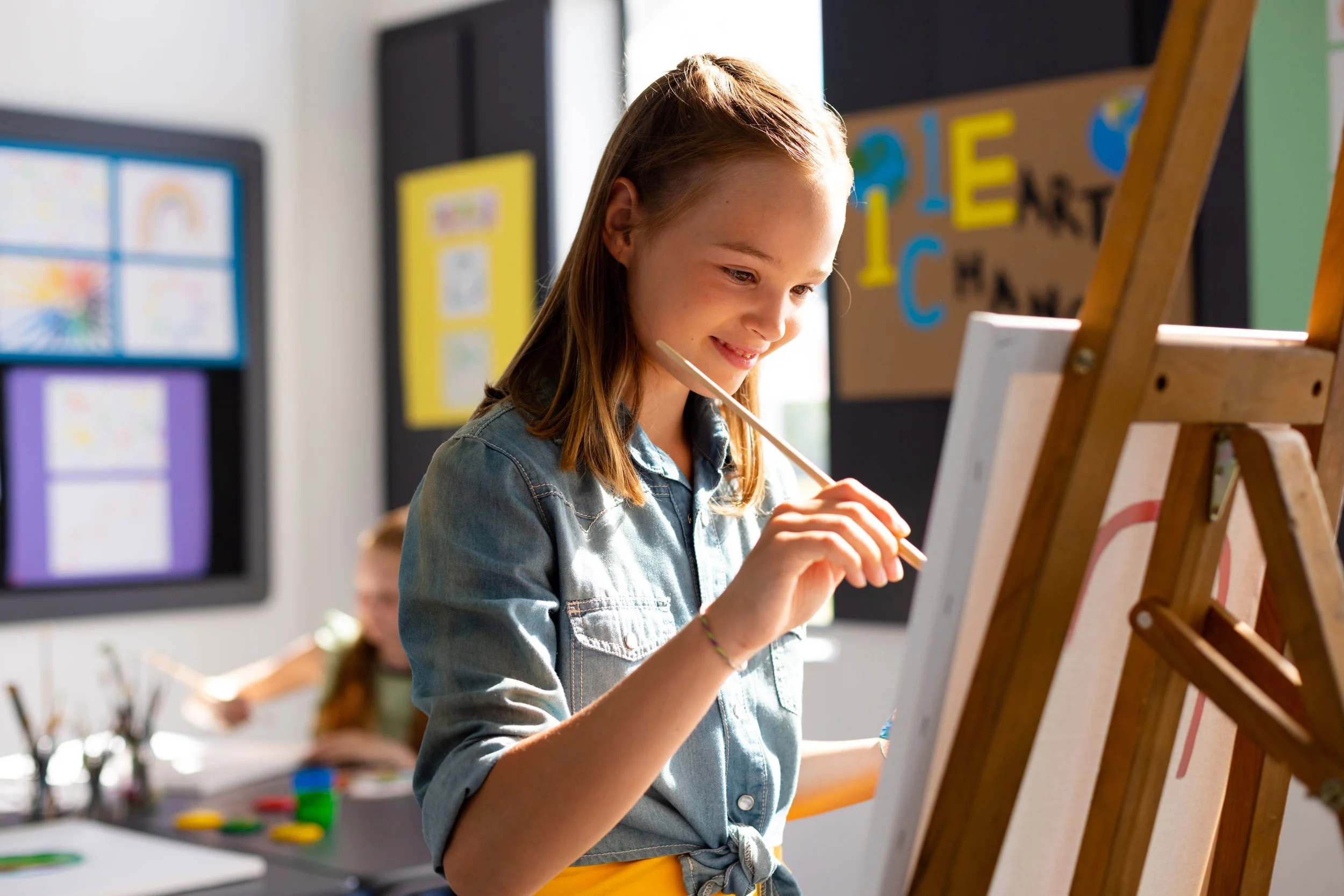 Young girl painting on a canvas in an art classroom with colorful posters on the walls.