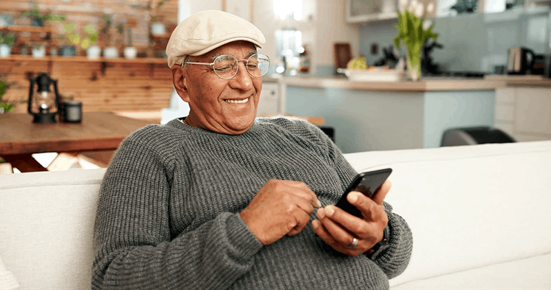 An elderly man wearing a beige cap, glasses, and a gray sweater, smiling and using his smartphone while sitting on a white couch in a cozy, well-lit kitchen and living area.