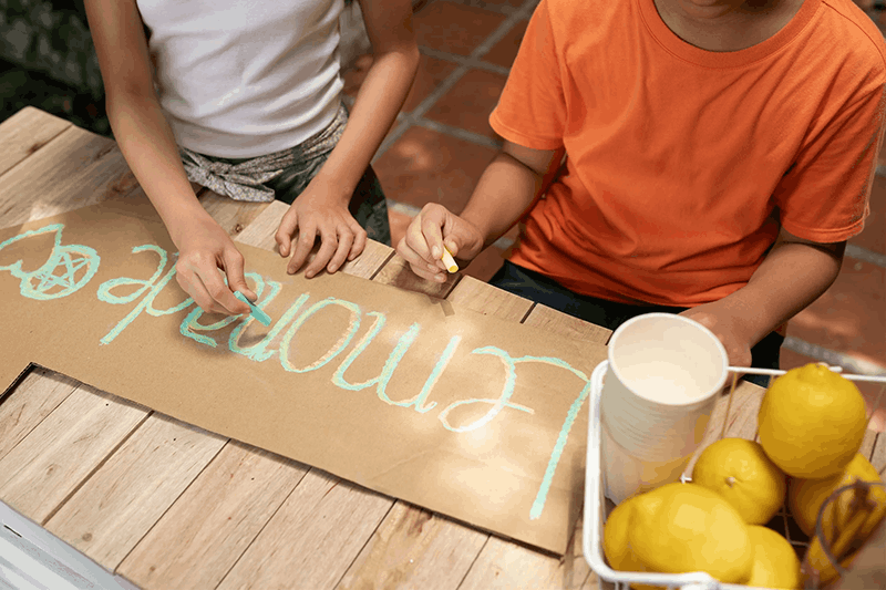 Two children drawing on a cardboard sign that says 'Welcome' using pastel chalks at an outdoor table with lemons and cups nearby.