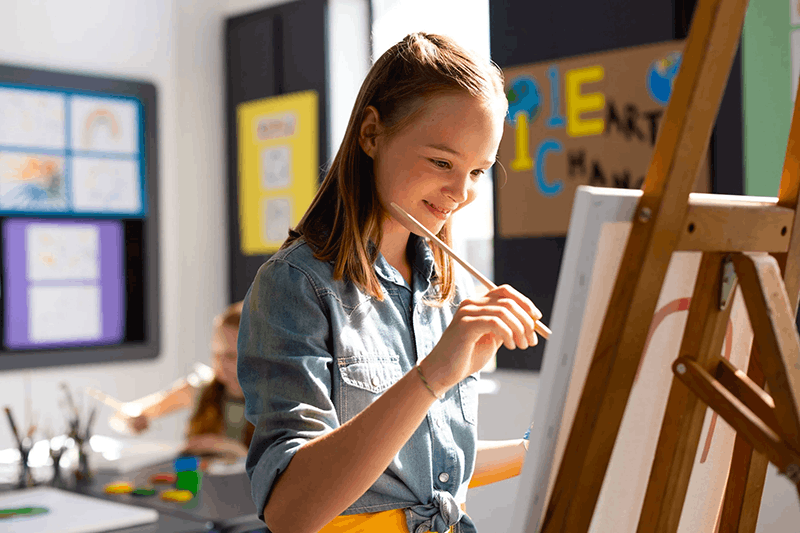 Young girl painting on a canvas in an art classroom with colorful posters on the walls.