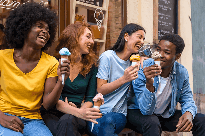 Four friends sitting on a bench outside an ice cream shop, laughing and enjoying ice cream cones.