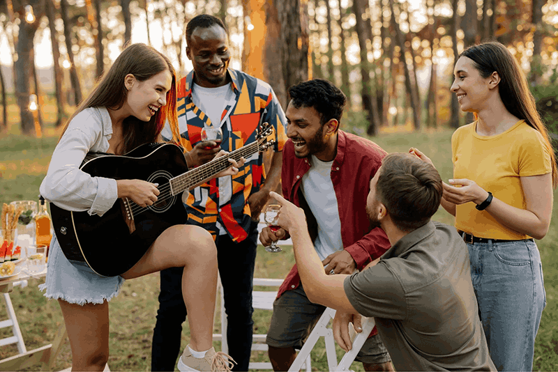 Group of friends enjoying a summer outdoor gathering, with one woman playing guitar and singing, others laughing and chatting, in a park with trees and string lights.