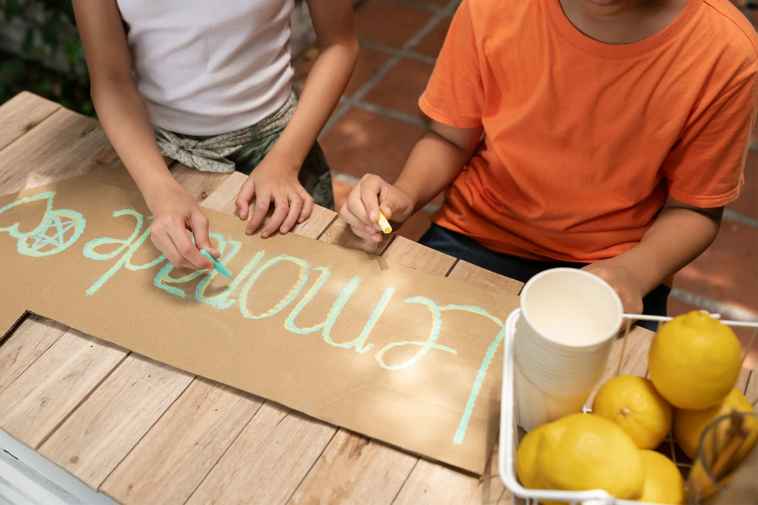 Two children drawing on a cardboard sign that says 'Welcome' using pastel chalks at an outdoor table with lemons and cups nearby.