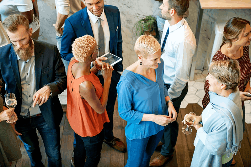 People having a social gathering at an indoor event, engaging in conversation, some holding glasses of white wine, with a mix of men and women dressed in business casual and semi-formal attire.