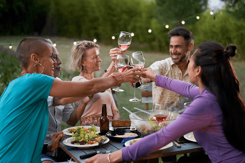 Group of six people celebrating outdoors, clinking wine glasses filled with rosé wine, surrounded by food and salad on a table, with string lights and trees in the background.