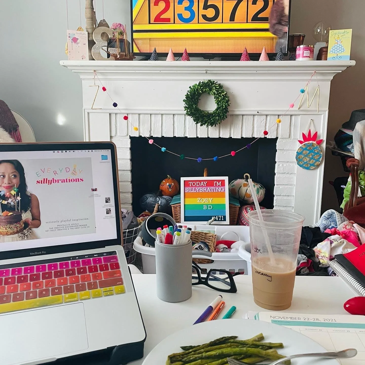 A decorated fireplace mantel with a small wreath, party hats, and a pineapple-shaped decoration. A lit birthday candle and a sign that reads 'Today I'm sillybrating Zoey B D' are in front of the fireplace. A computer screen, markers, glasses, a drink, and a plate of cooked asparagus are on the desk in front. Decorations and supplies indicate a birthday celebration.