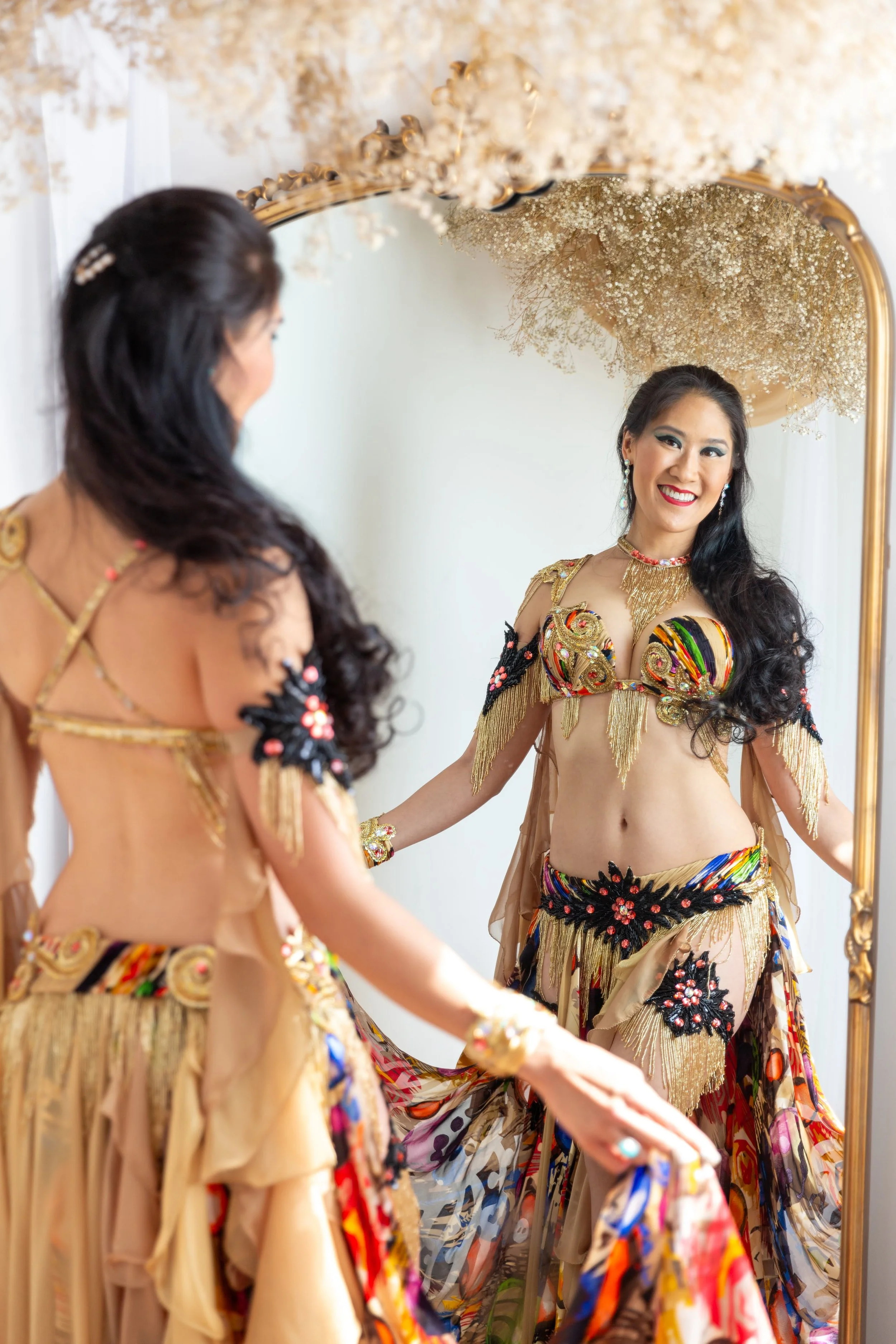 stephaniebellydance.com features Stephanie in a Turlock studio photoshoot wearing a cream and multi-color costume smiling at her reflection in a gold mirror under a cloud of dried white baby's breath flowers.