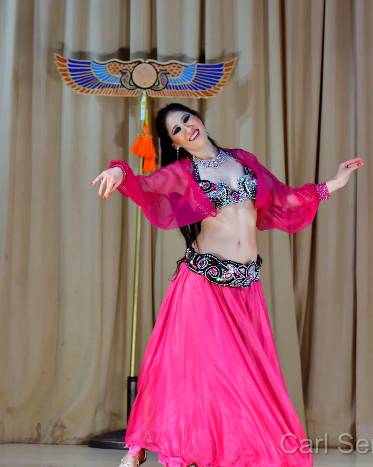 Stephanie performing a belly dance on stage, wearing a pink skirt and black and silver beaded top, in front of beige curtains with ornamental props.