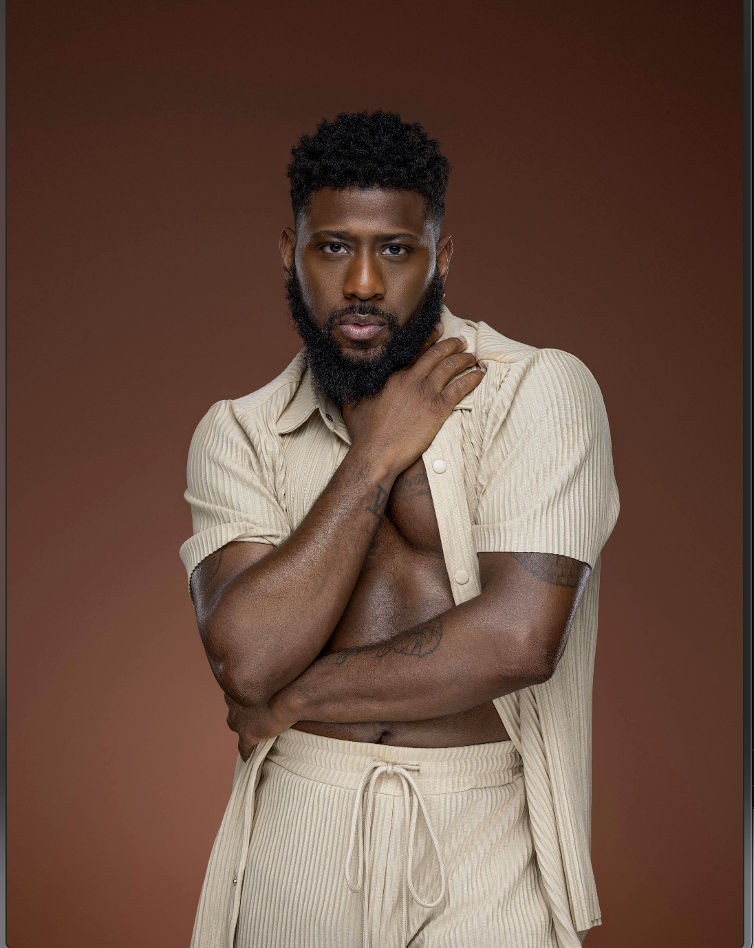A young man with a beard and tattoos on his arm, wearing a beige short-sleeved shirt and matching pants, poses against a brown background, touching his neck with one hand and crossing his other arm.