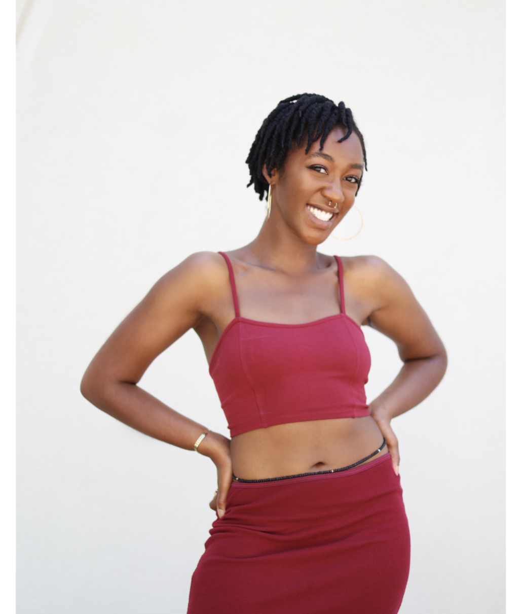 Woman with short dreadlocks, gold hoop earring, and nose piercing smiling confidently, wearing a red crop top and matching skirt.