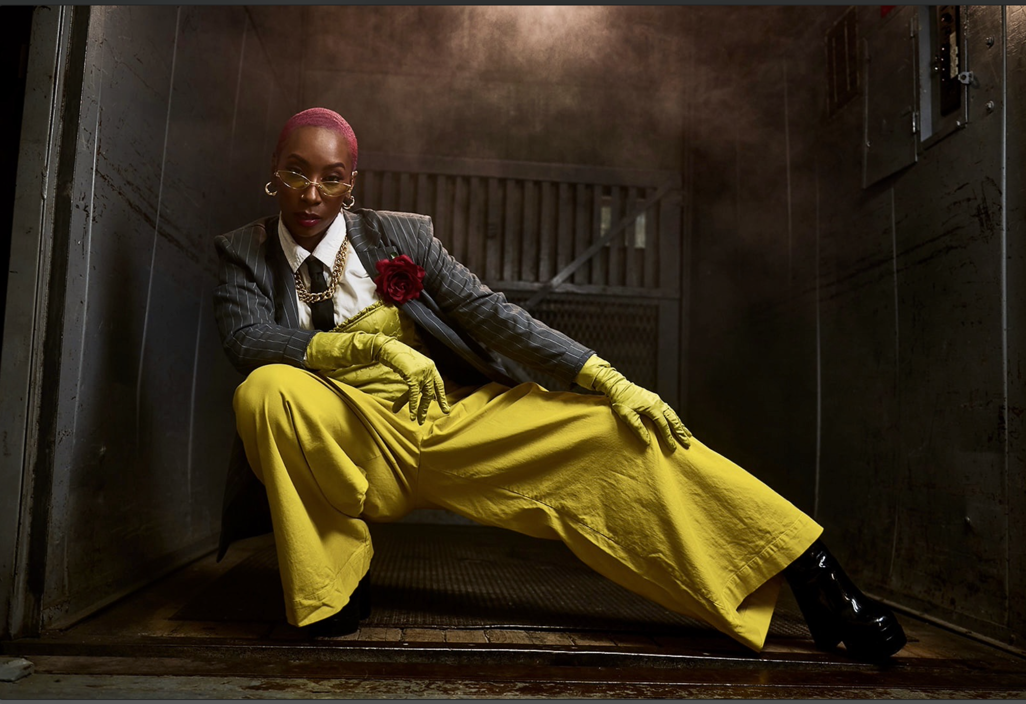 A person with pink hair, yellow-tinted sunglasses, a pinstripe suit jacket, a white shirt, a black tie, large earrings, a gold chain necklace, a red rose brooch, yellow gloves, bright yellow wide-leg pants, and black boots, sitting in a metal elevator shaft with industrial background, posing confidently.