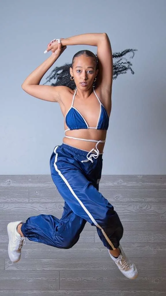 Woman in athletic attire jumping with one arm raised and the other across her head against a plain gray background.