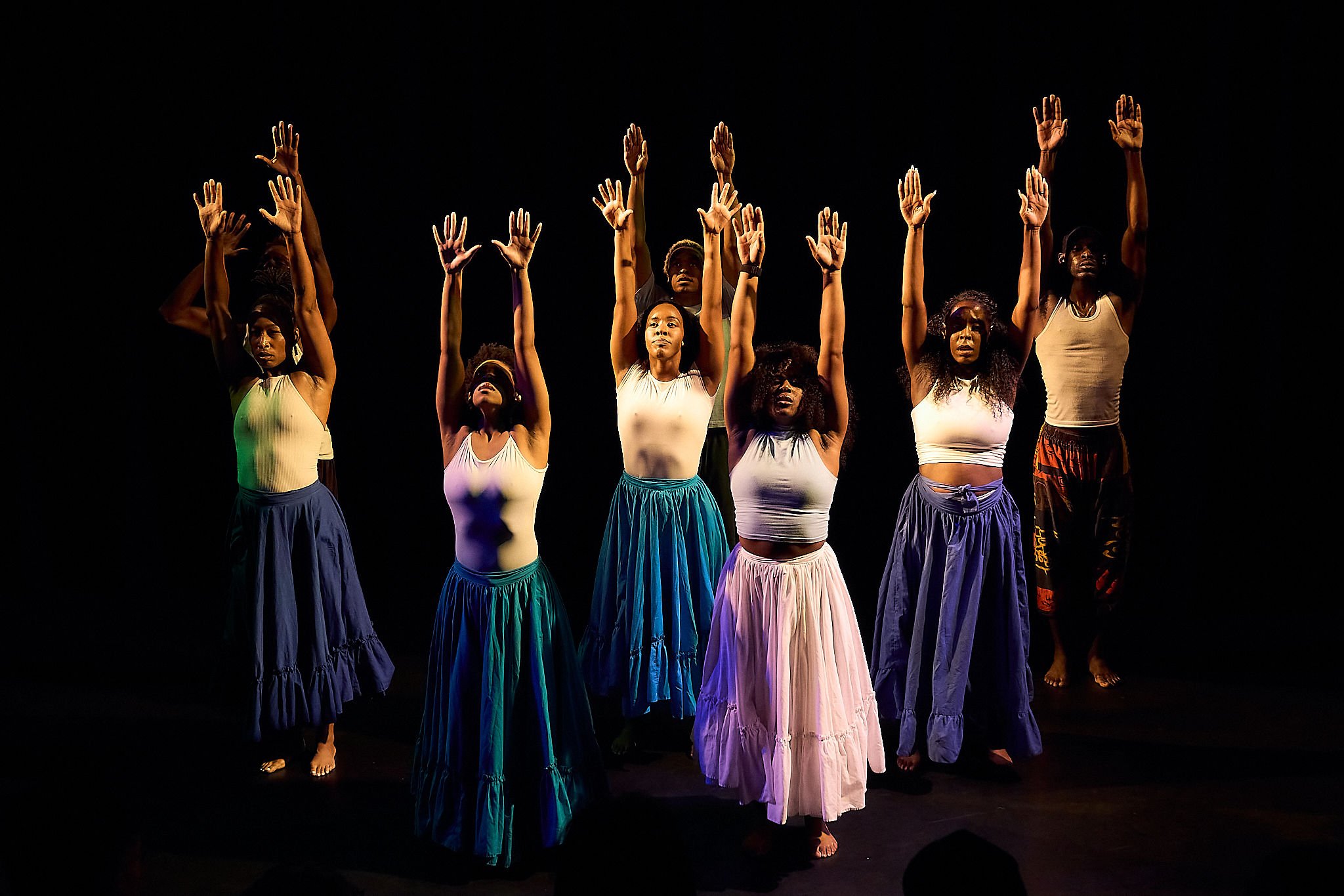 Group of six women dancing with arms raised, wearing long skirts and tank tops on a dark stage.