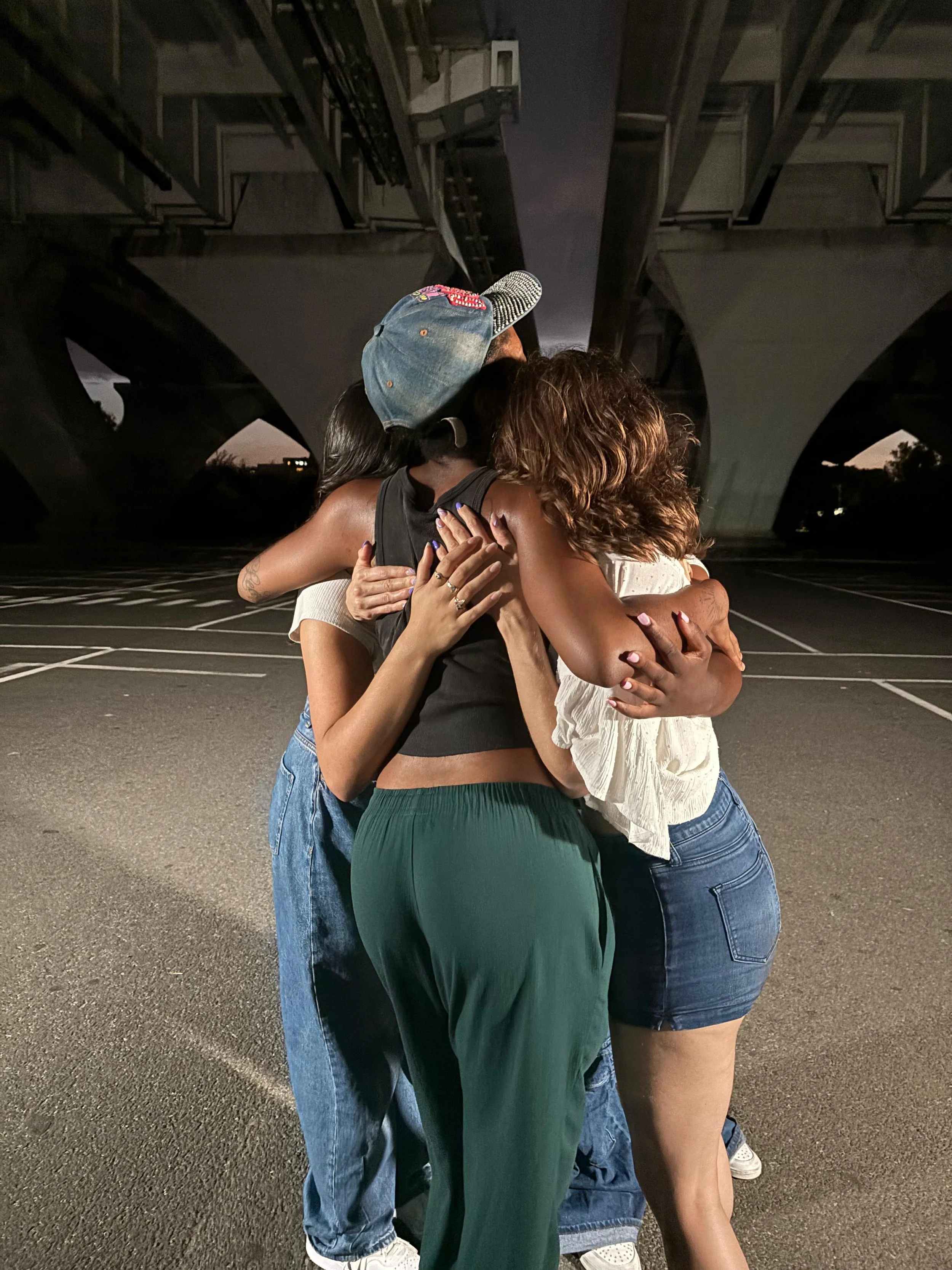 Four women hugging under a bridge in an empty parking lot at dusk.