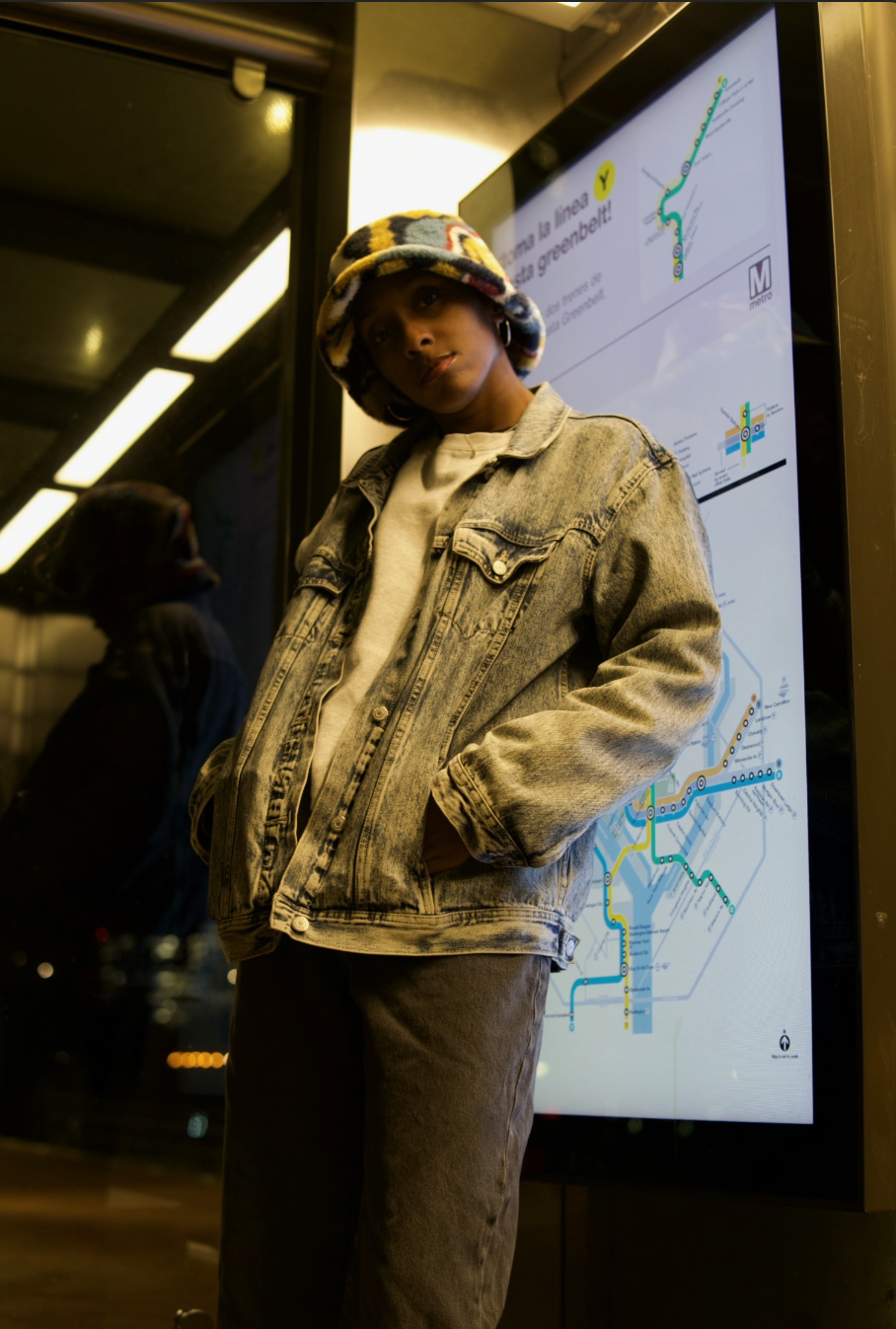 A woman standing in front of a digital map display at a subway station, wearing a colorful bucket hat, denim jacket, and casual clothing.
