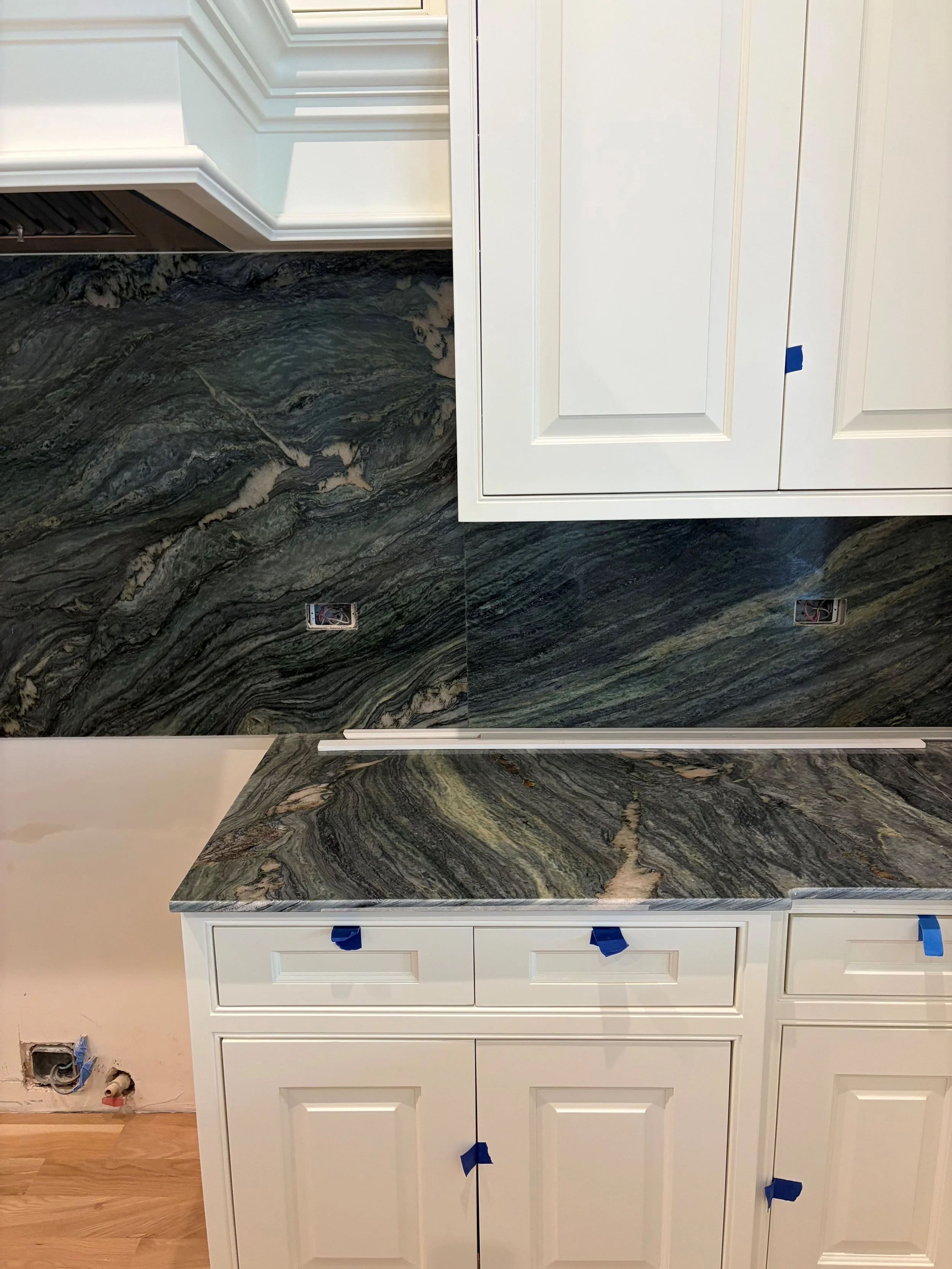 Kitchen cabinets with a dark marble countertop and backsplash, white cabinet doors, blue painter's tape, and unfinished electrical outlets.