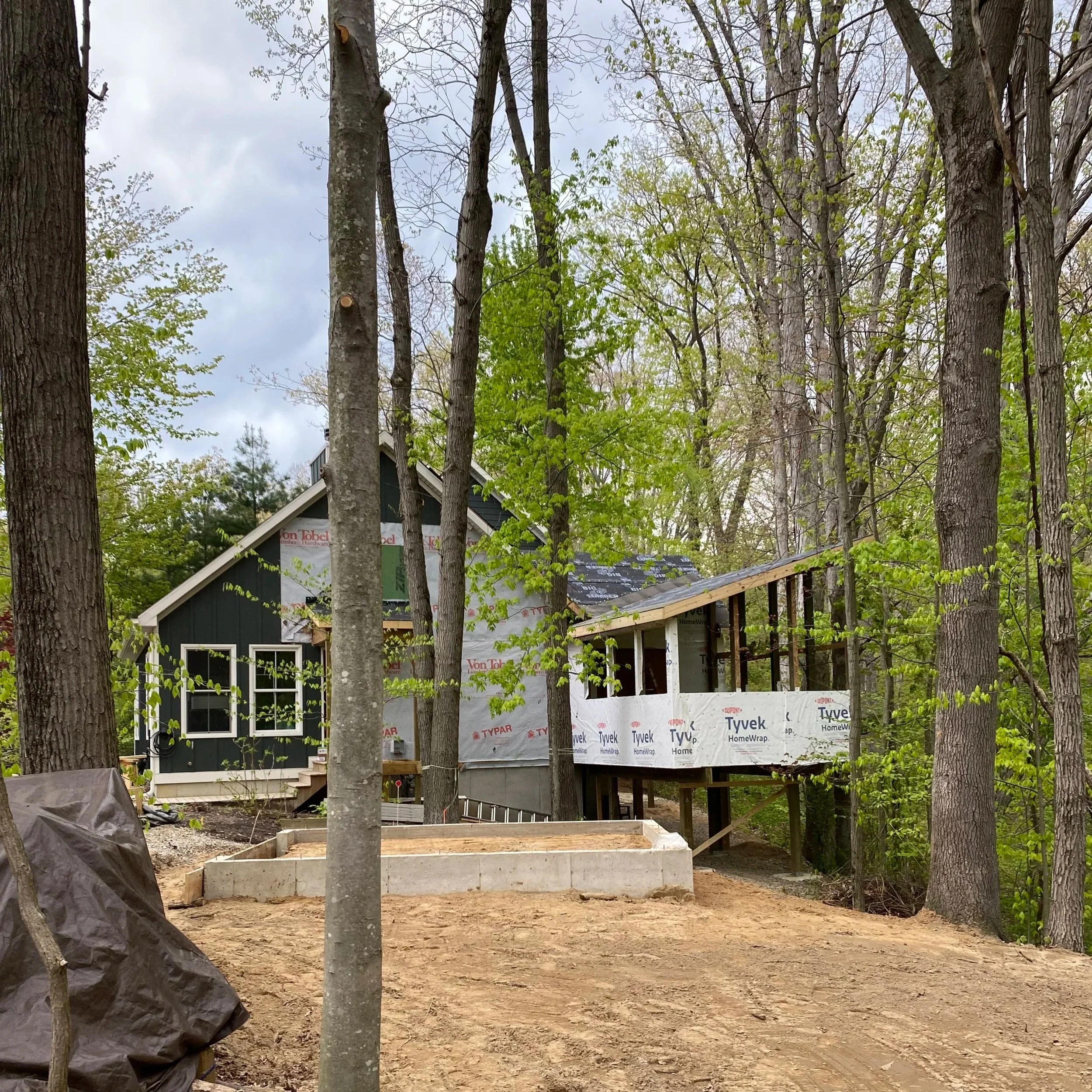 A house under construction in a wooded area with trees and a cloudy sky in the background. The house has black siding, white trim, and a partially finished roof with visible roofing materials.
