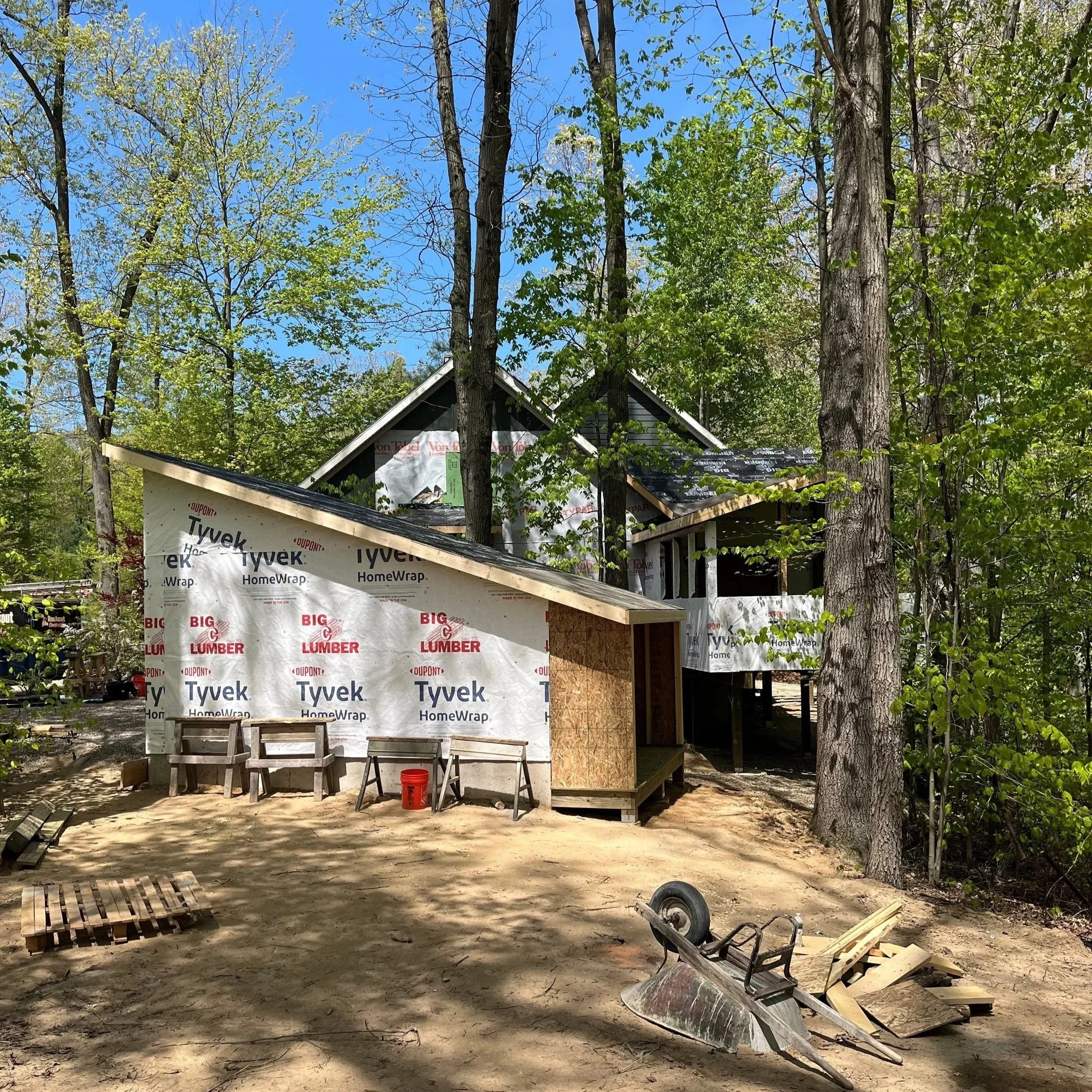 A house under construction surrounded by trees with green leaves and a clear blue sky. The house has black roofing with some building materials and tools in the front yard.