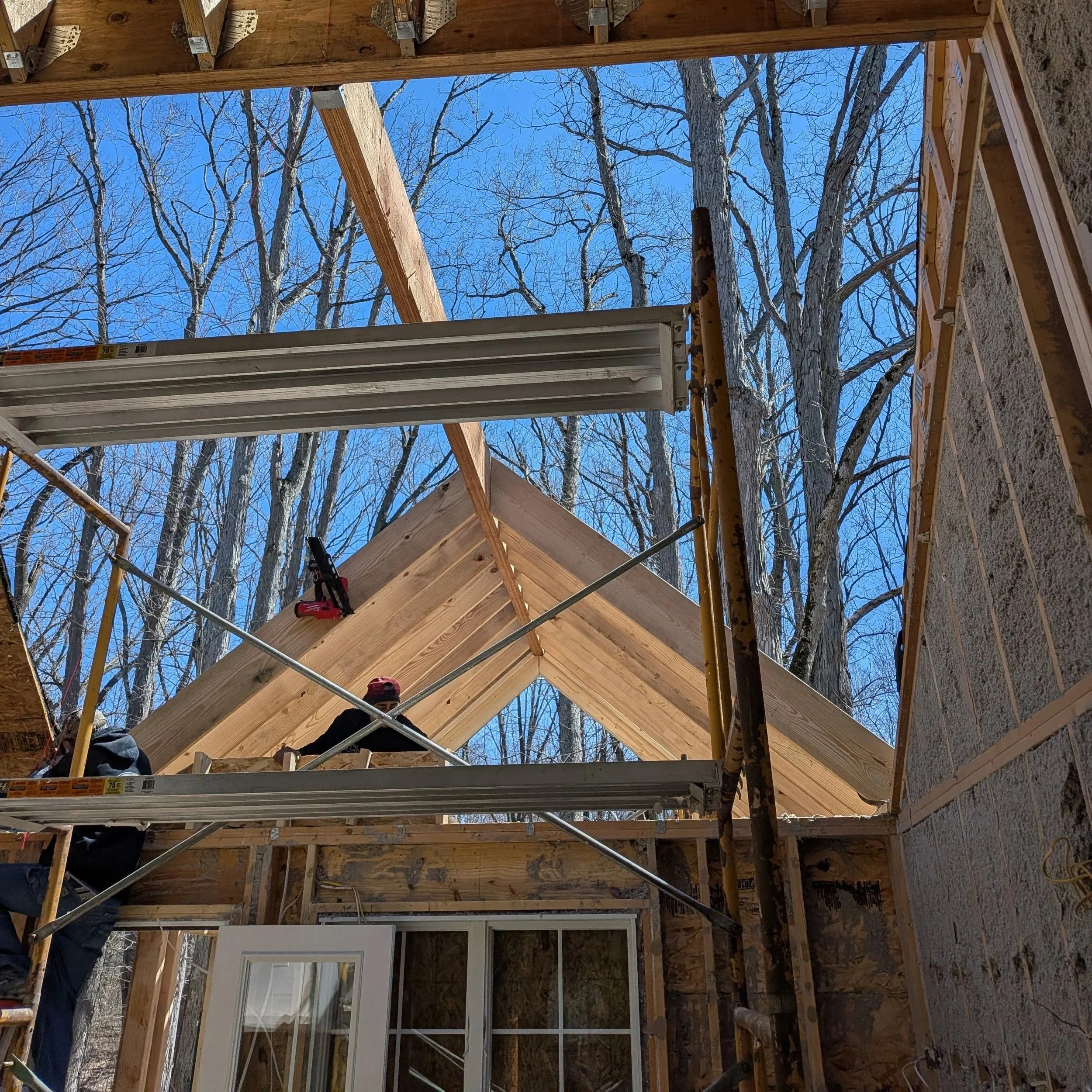 Construction site with wooden framing, scaffolding, and a large window, with leafless trees in the background.