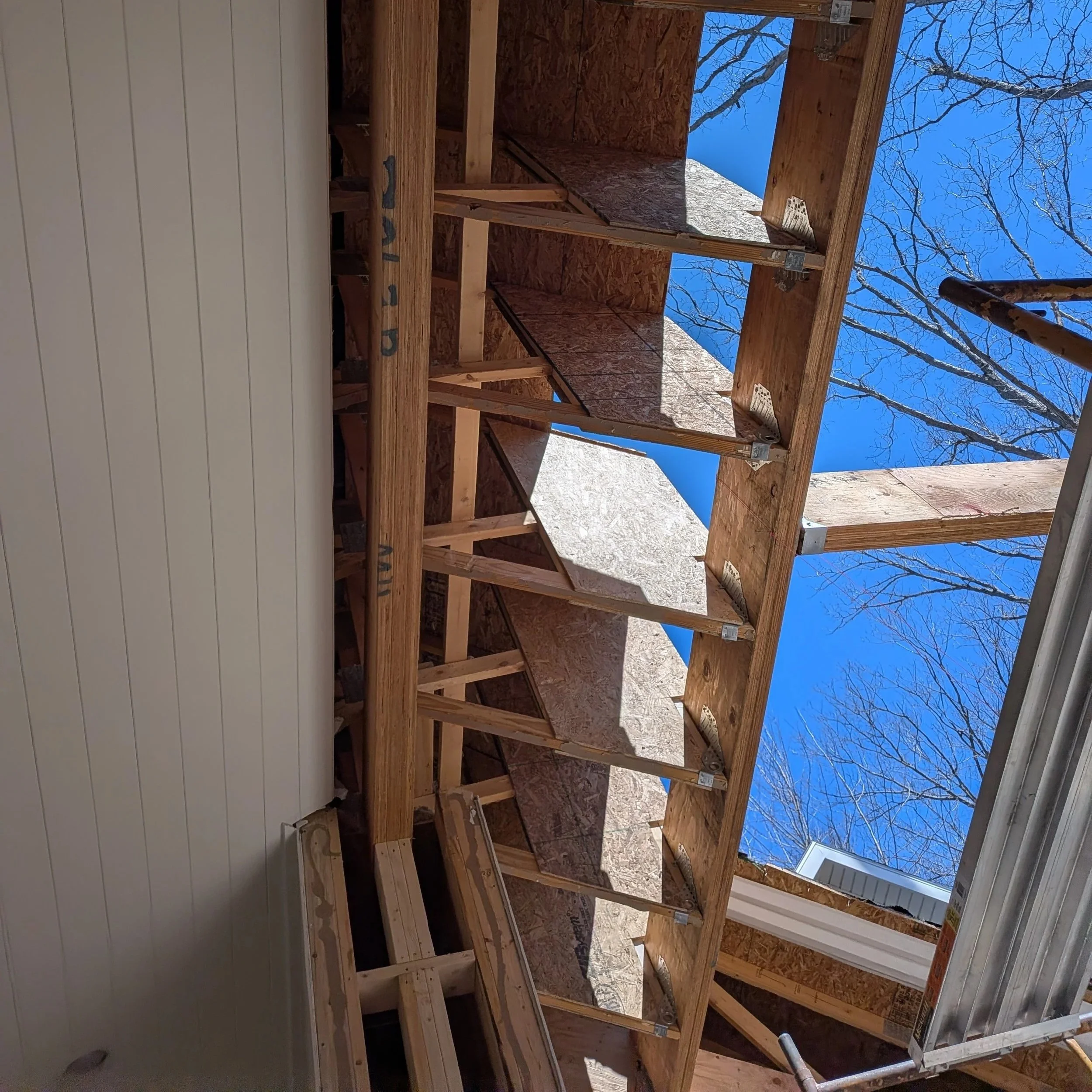 Construction site with wooden framing for building walls, blue sky and leafless trees visible in the background.