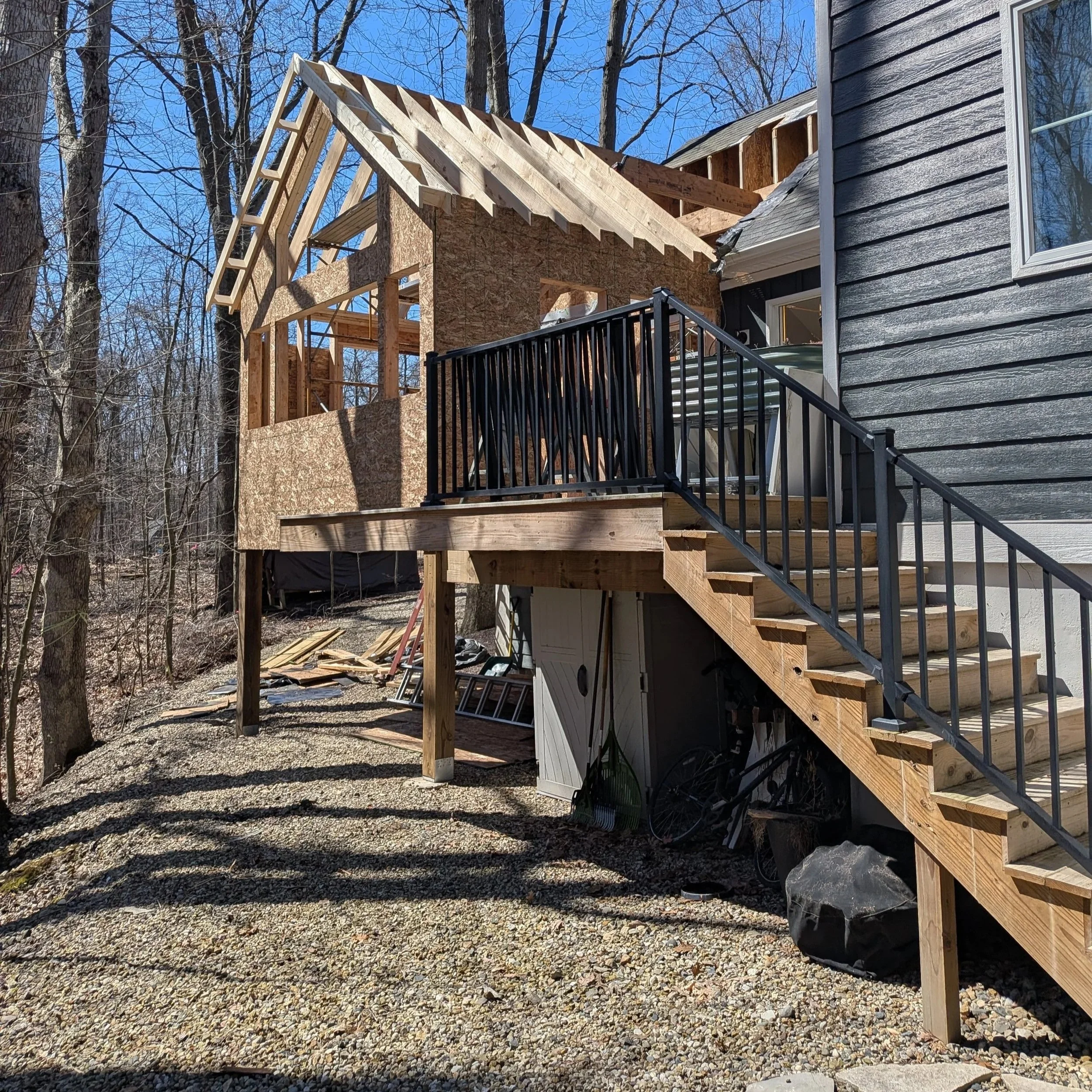 House under construction with framed roof and black railing staircase, surrounded by leafless trees on a gravel yard in a sunny, clear day.